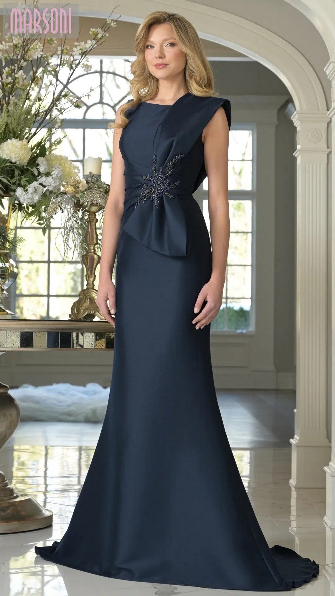 A woman stands indoors near a floral arrangement, wearing the Marsoni MV1359 long formal dress in navy. The sleeveless gown features a fitted waist and decorative embellishment, exuding elegant sophistication.